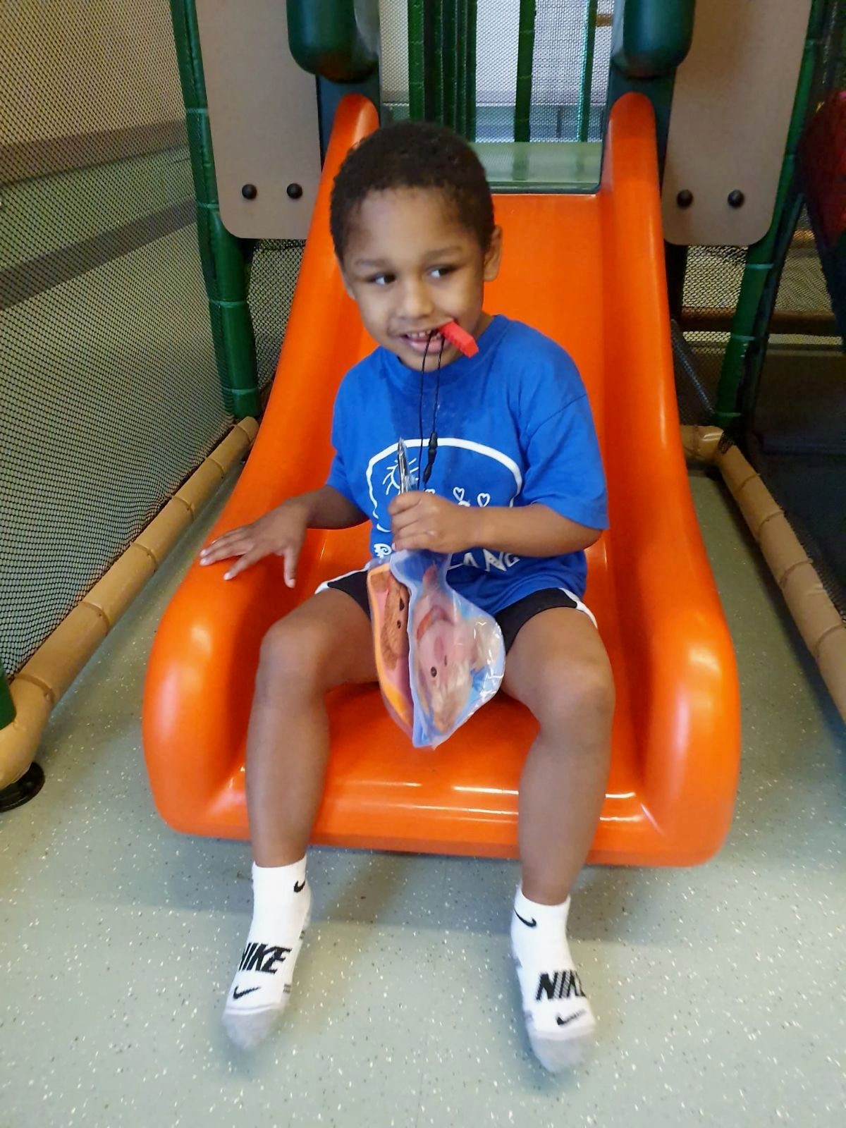 young smiling child wearing a blue t-shirt and dark blue shorts sits on an orange slide. He’s holding a plastic bag in his left hand and has a whistle in his mouth.