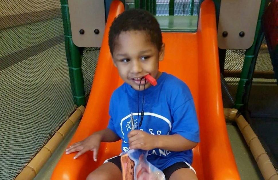 Un jeune enfant souriant, vêtu d’un t-shirt bleu et de culottes courtes bleu foncé, est assis sur une glissade orange. Il tient un sac en plastique dans la main gauche et a un sifflet dans la bouche. 
