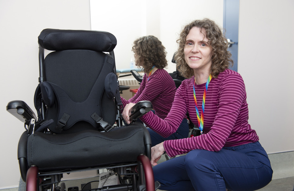 A woman with brown hair and a pink striped shirt poses beside a wheelchair