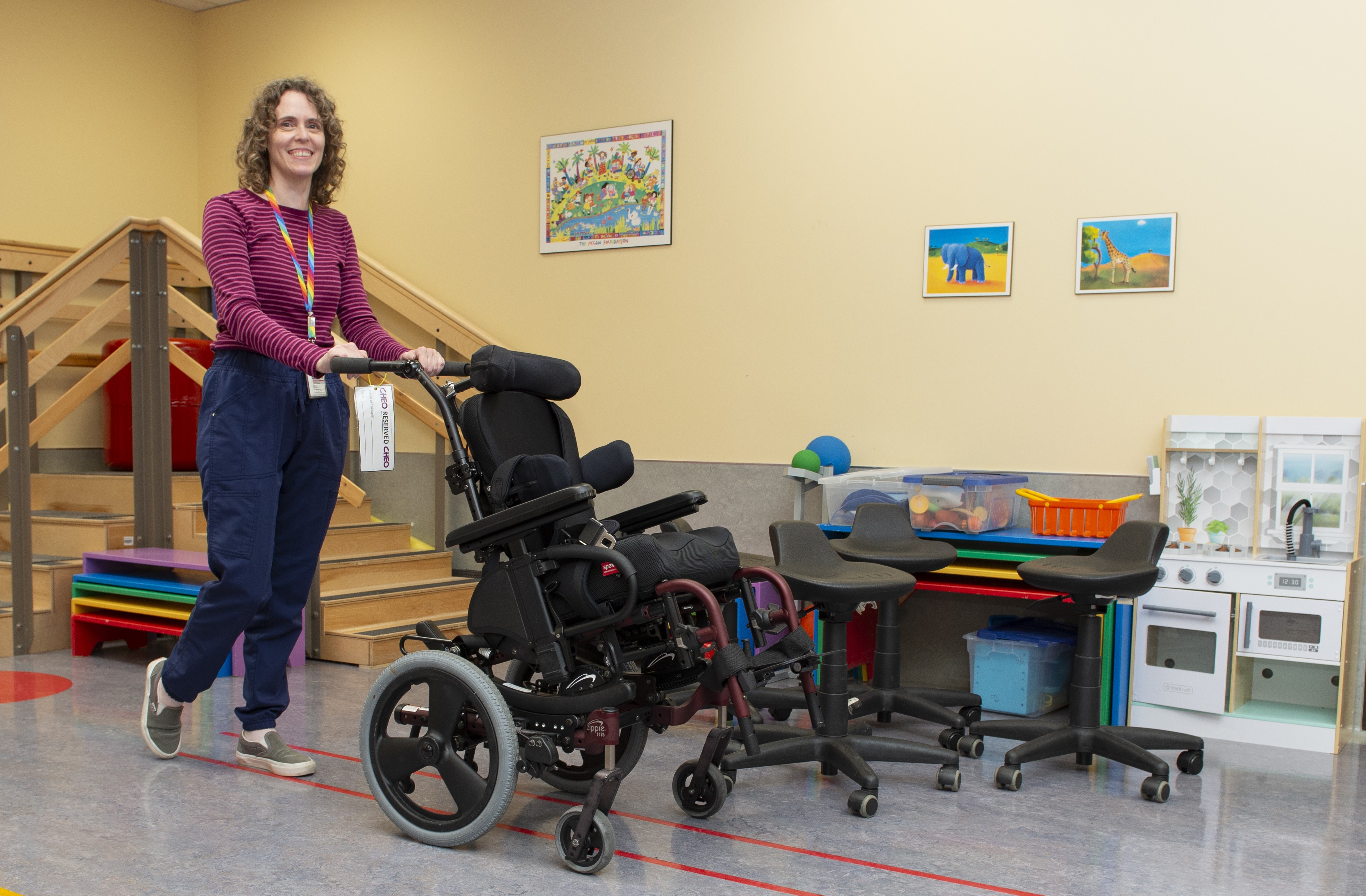 A woman with brown hair and a pink shirt pushes a wheelchair while smiling at the camera