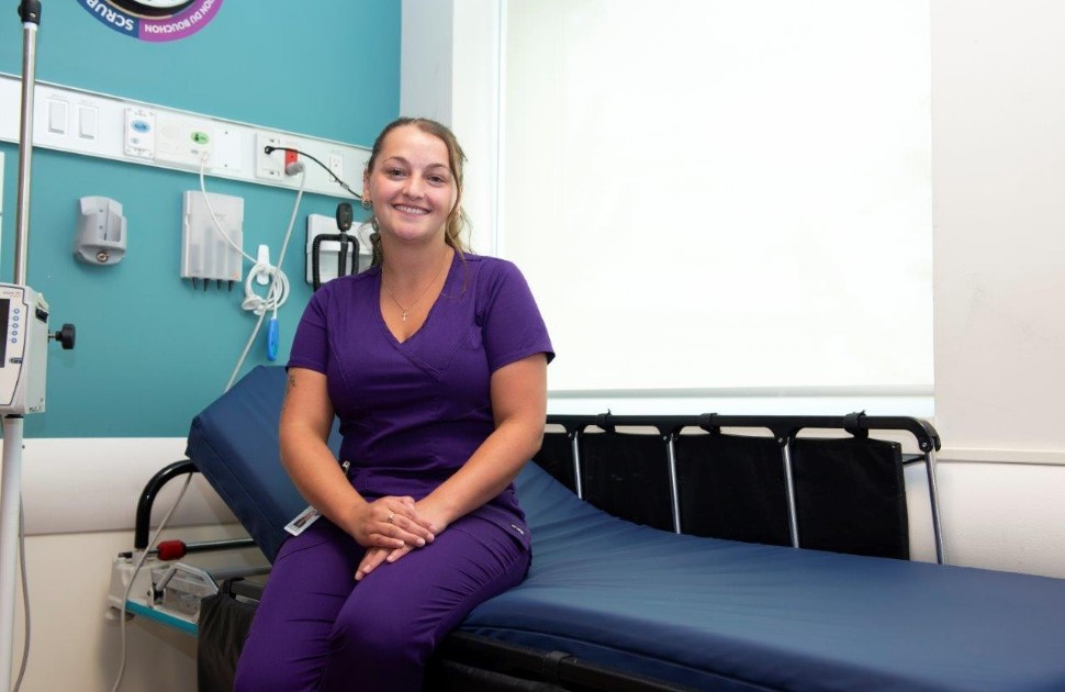 Isabelle Moss. Elle porte une blouse bleu foncé et est assise sur un brancard d'hôpital.