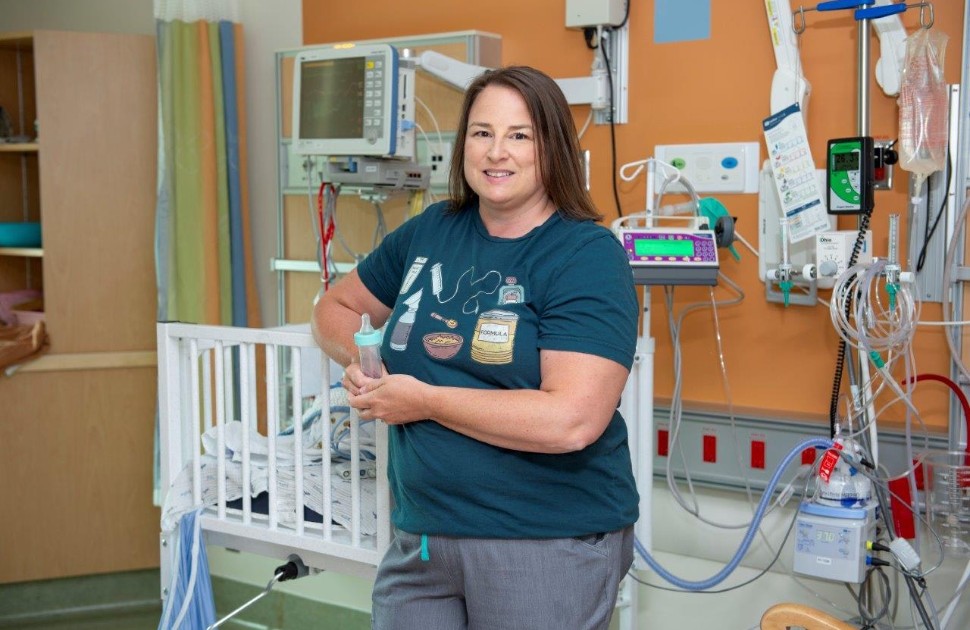 Jeanette wears a green graphic t-shirt and stands in front a NICU crib. She holds a feeding bottle.