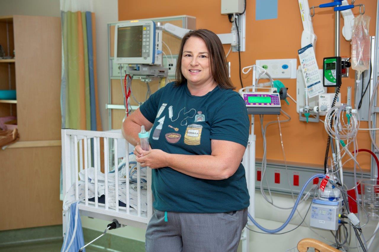 Jeanette wears a green graphic t-shirt and stands in front a NICU crib. She holds a feeding bottle.