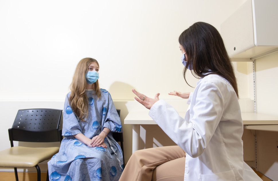 A woman with brown hair and a white lab coat speaks to a patient with blonde hair in a blue down