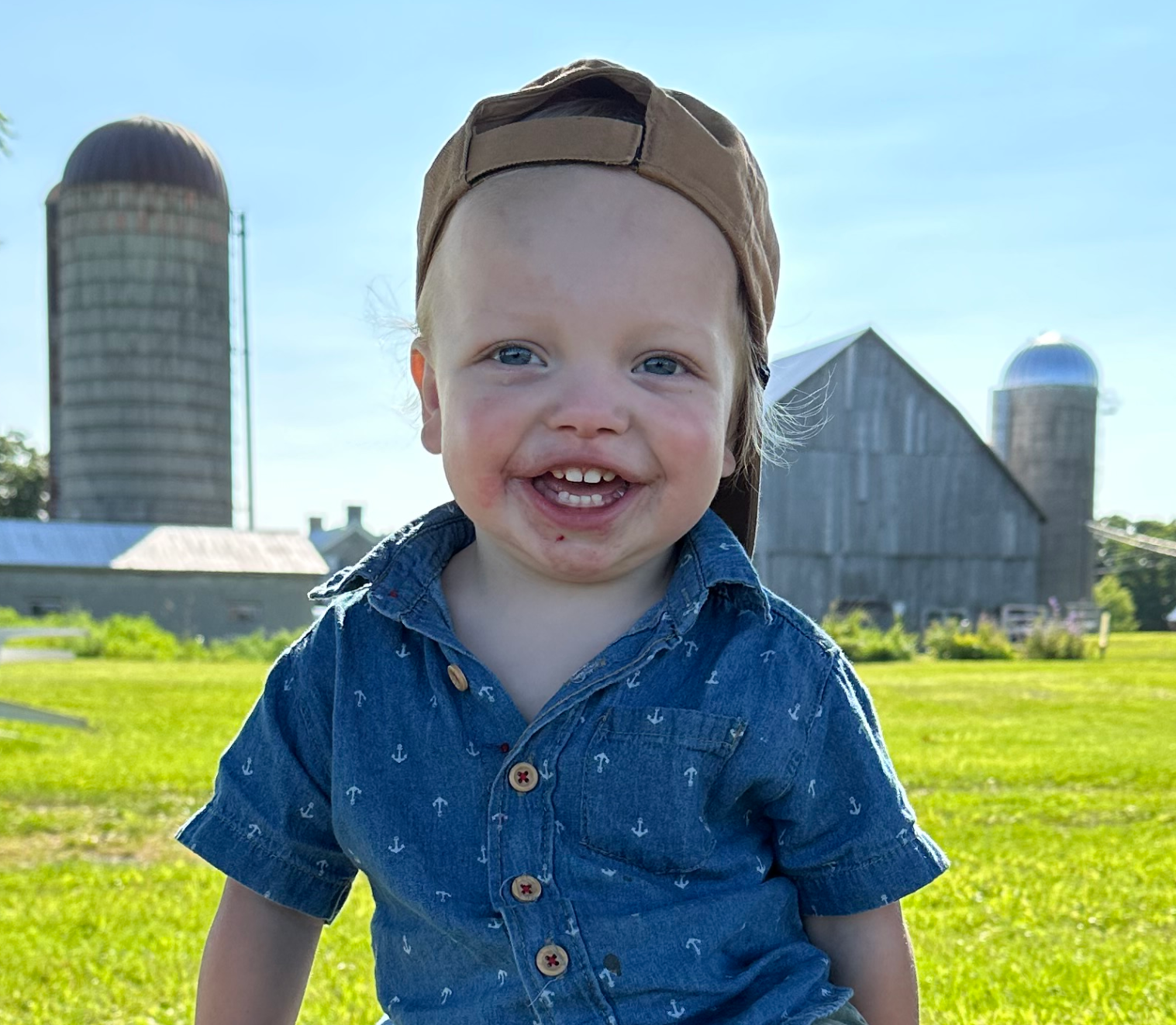 boy smiles while sitting on tree stump in front of farm in summer weather